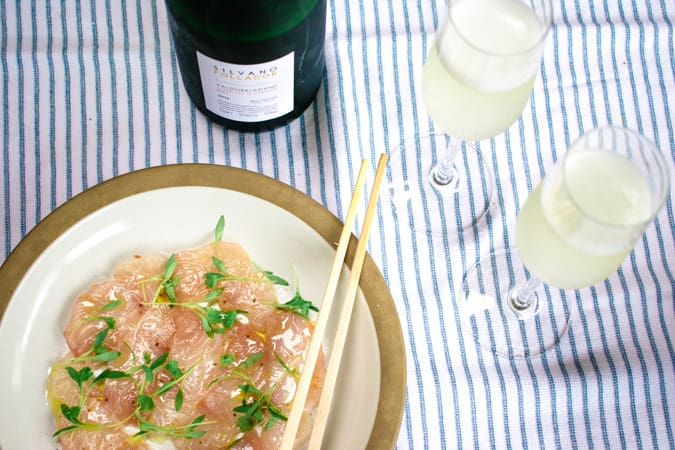Plate of yellowtail crudo with chopsticks and 2 glasses and bottle of prosecco wine on a blue and white striped tablecloth.