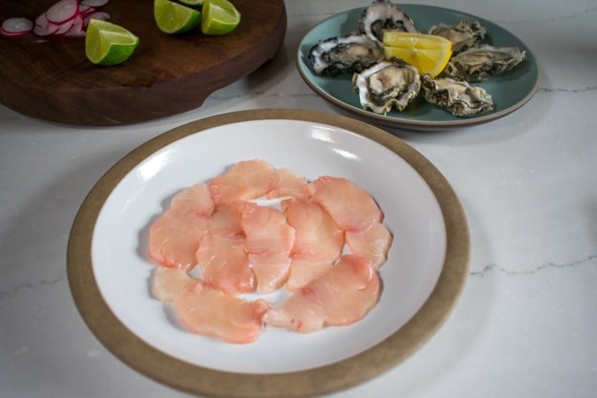 Slices of raw yellowtail crudo arranged on a white plate with oysters in the background.