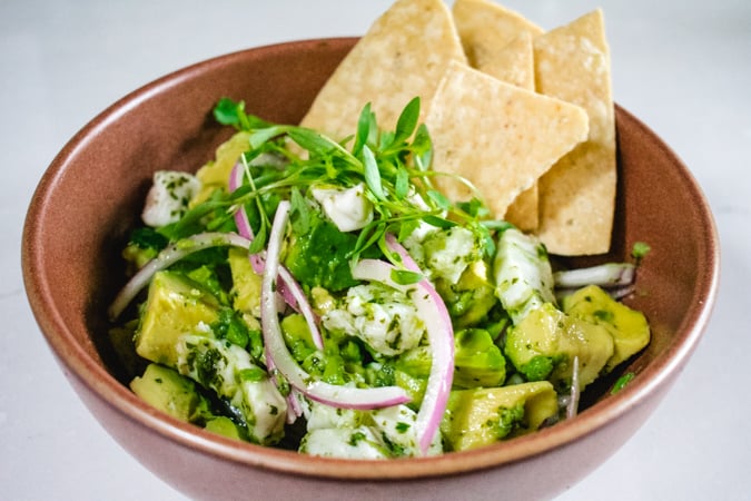 Brown bowl of halibut ceviche with avocado, red onion, micro cilantro, and tortilla chip garnish.