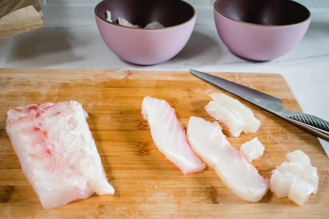 Halibut sliced into dice on cutting board with knife and 2 pink bowls in the background.