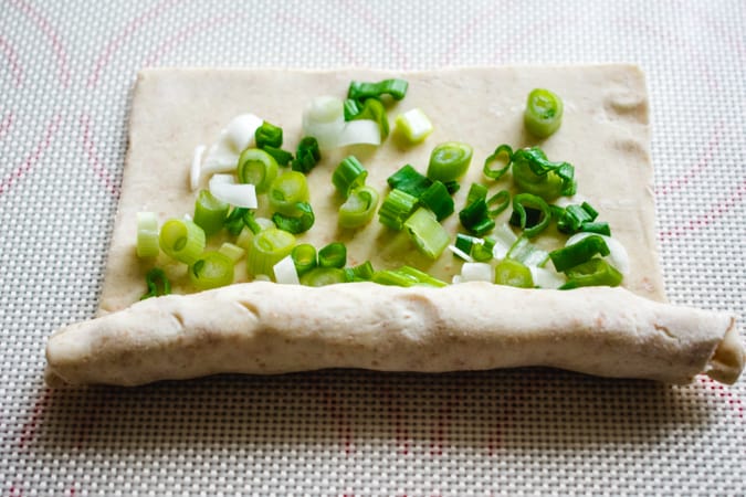 Half rolled up square of puff pastry with chopped scallions on a silicone baking mat