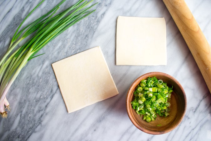 Puff pastry squares, green onion, chopped green onion, and wooden rolling pin on a marble board