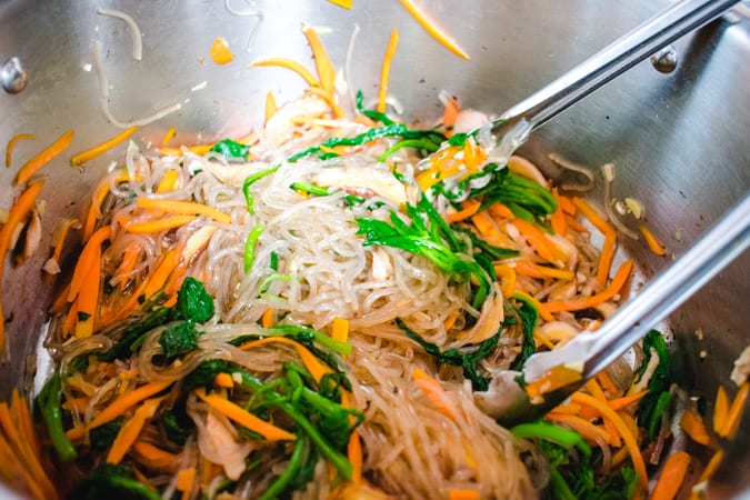 Korean japchae being stirred in a silver pot with tongs.