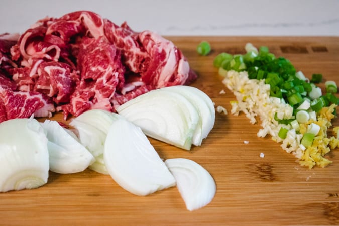 sliced onion, beef, garlic, ginger, and green onion on a wood cutting board