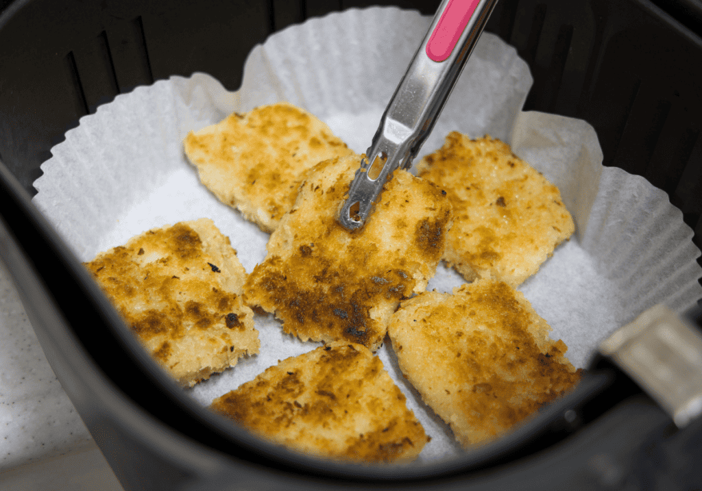 breaded fish nuggets with tongs and a parchment paper liner in an air fryer basket