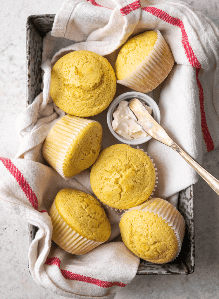 Corn muffins in a baking pan with a red and white towel and a ramekin of vegan butter with a knife