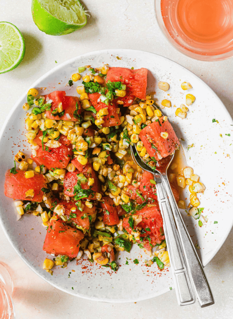 watermelon and charred corn salad on a white plate with a spoon and fork on the plate