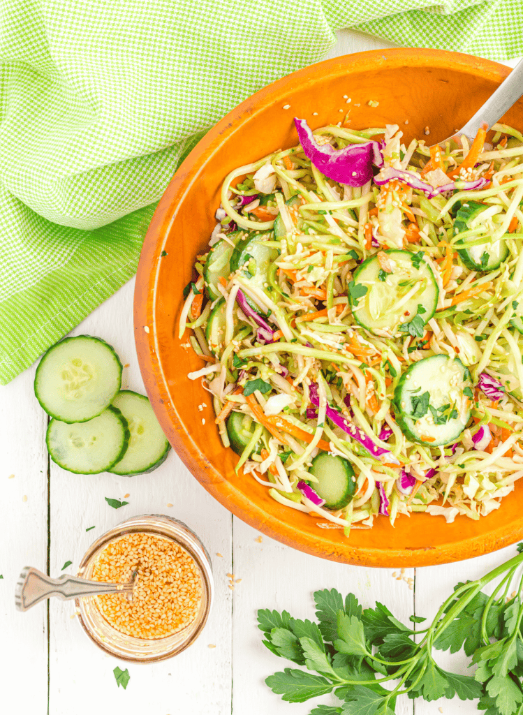 Broccoli slaw in an orange bowl with green towels and vegetables in the background