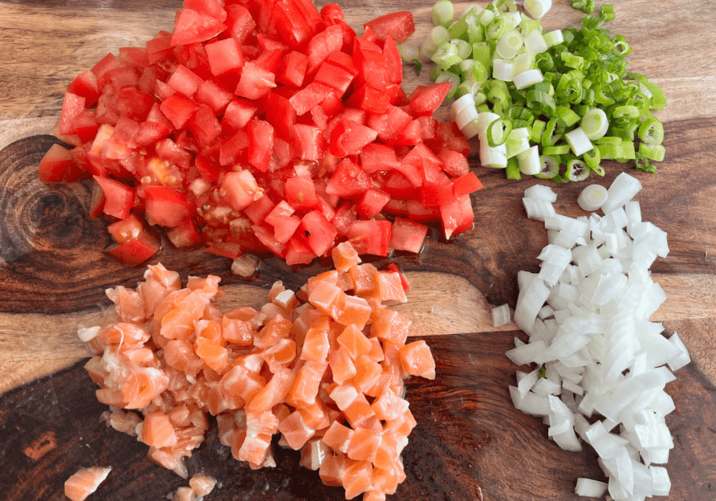 chopped tomatoes, salmon, green onion, and sweet onion on a wooden cutting board.