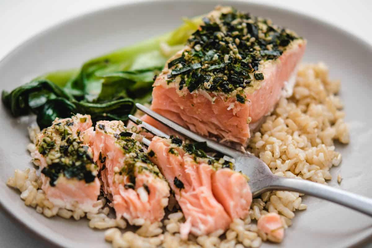 Filet of furikake salmon on a gray plate with brown rice, bok choy, and a fork with pieces of salmon in front of it.