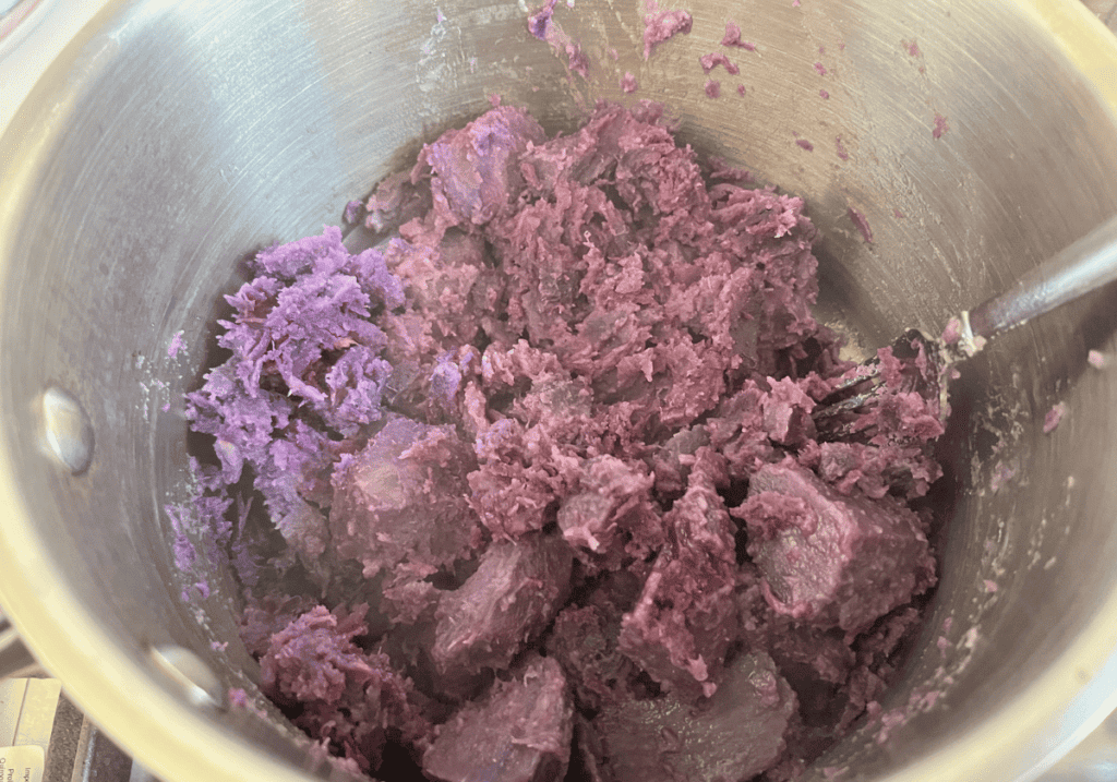 Chunks of purple potatoes being mashed with a fork in a silver saucepan.