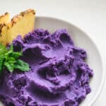 Baked purple potato mash in a white bowl with a garnish of pineapple wedges and mint.