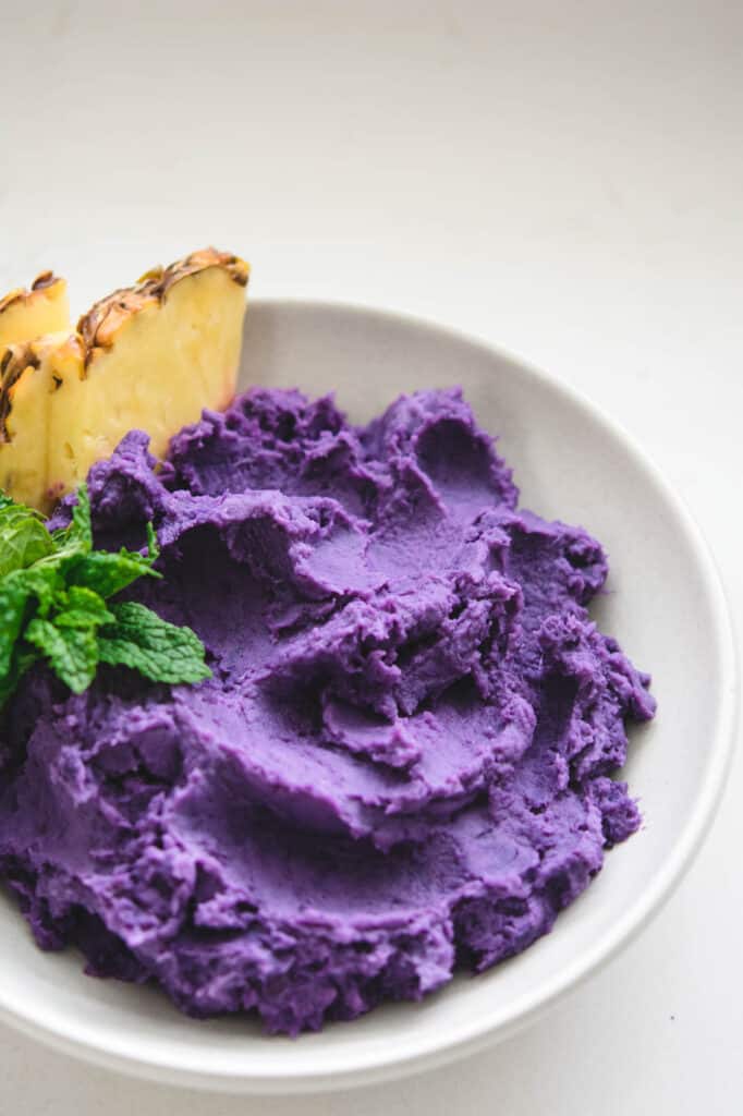 Baked purple potato mash in a white bowl with a garnish of pineapple wedges and mint.
