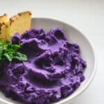 Purple potato mash in a white bowl with a garnish of pineapple wedges and mint.