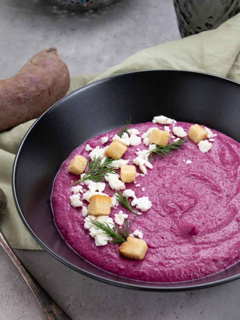 Purple sweet potato soup in a black bowl with a garnish of cheese crumbles, croutons, and fresh dill