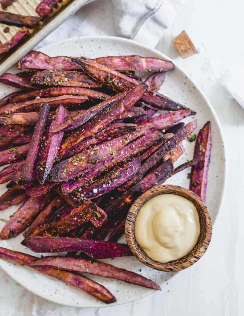 A mound of purple sweet potato fries on a white plate with a small brown bowl of mayo