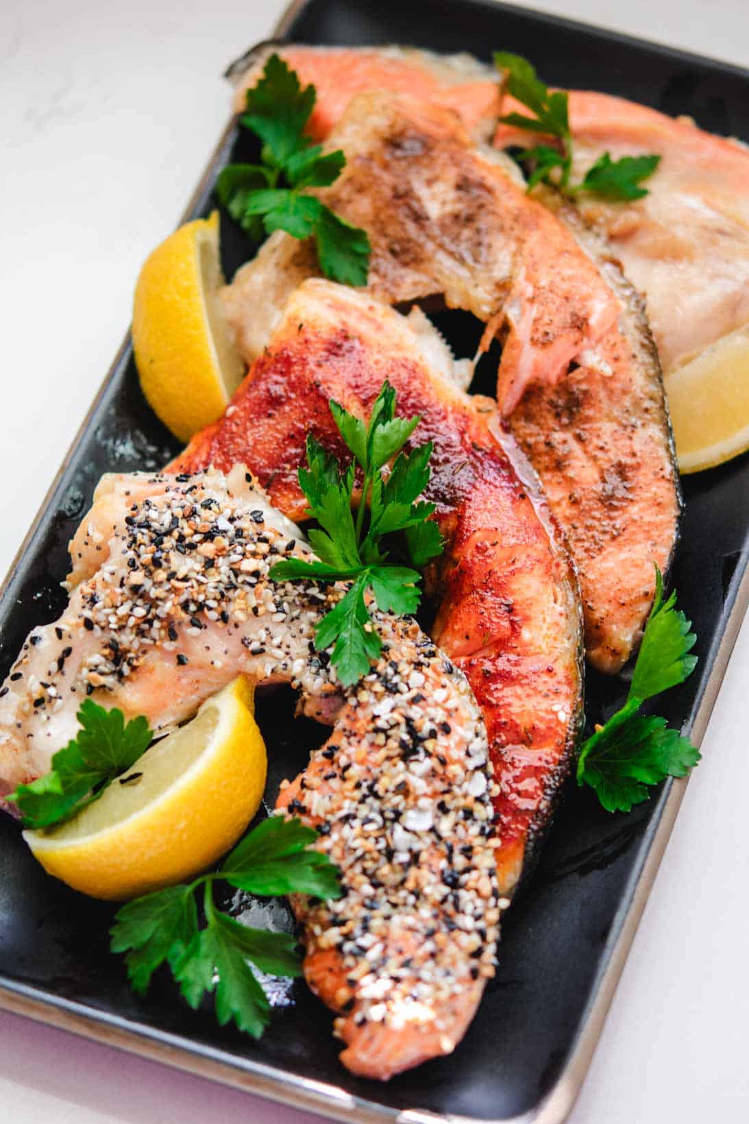 4 salmon collars with different toppings on a rectangular black plate with parsley garnish and lemon wedges