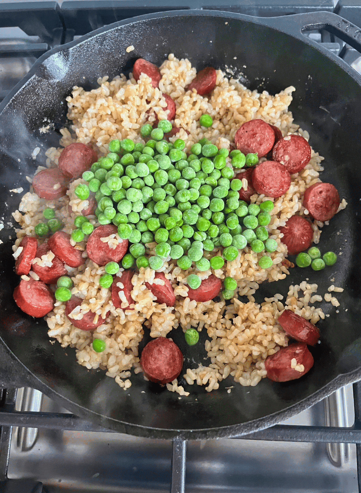frozen green peas on top of brown rice and hot dog slices in a cast iron pan