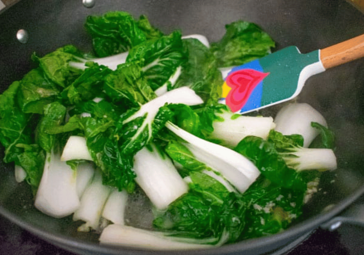 baby bok choy being stir fried in a black wok with a rubber spatula