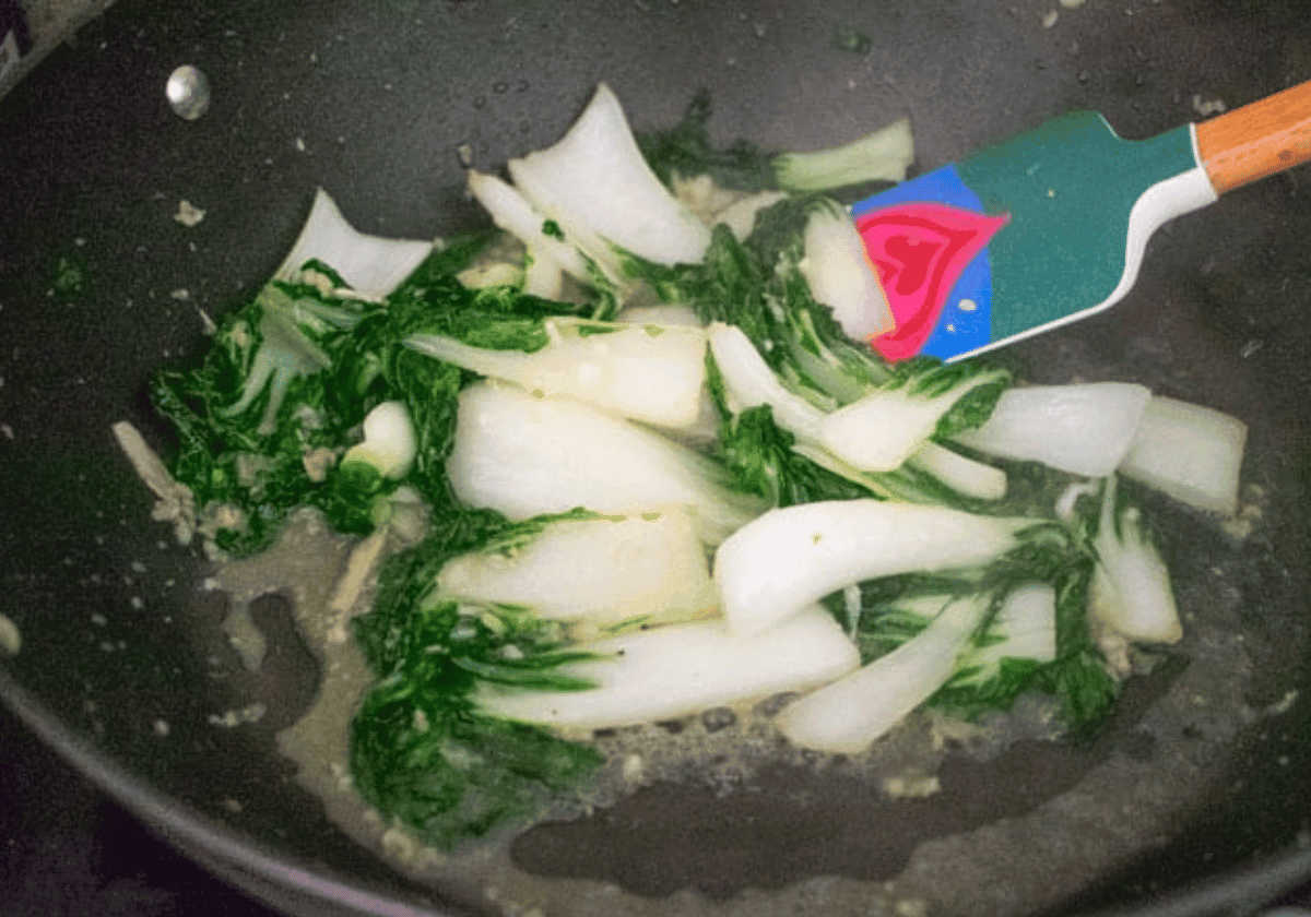 baby bok choy being stir fried in a black wok with a rubber spatula