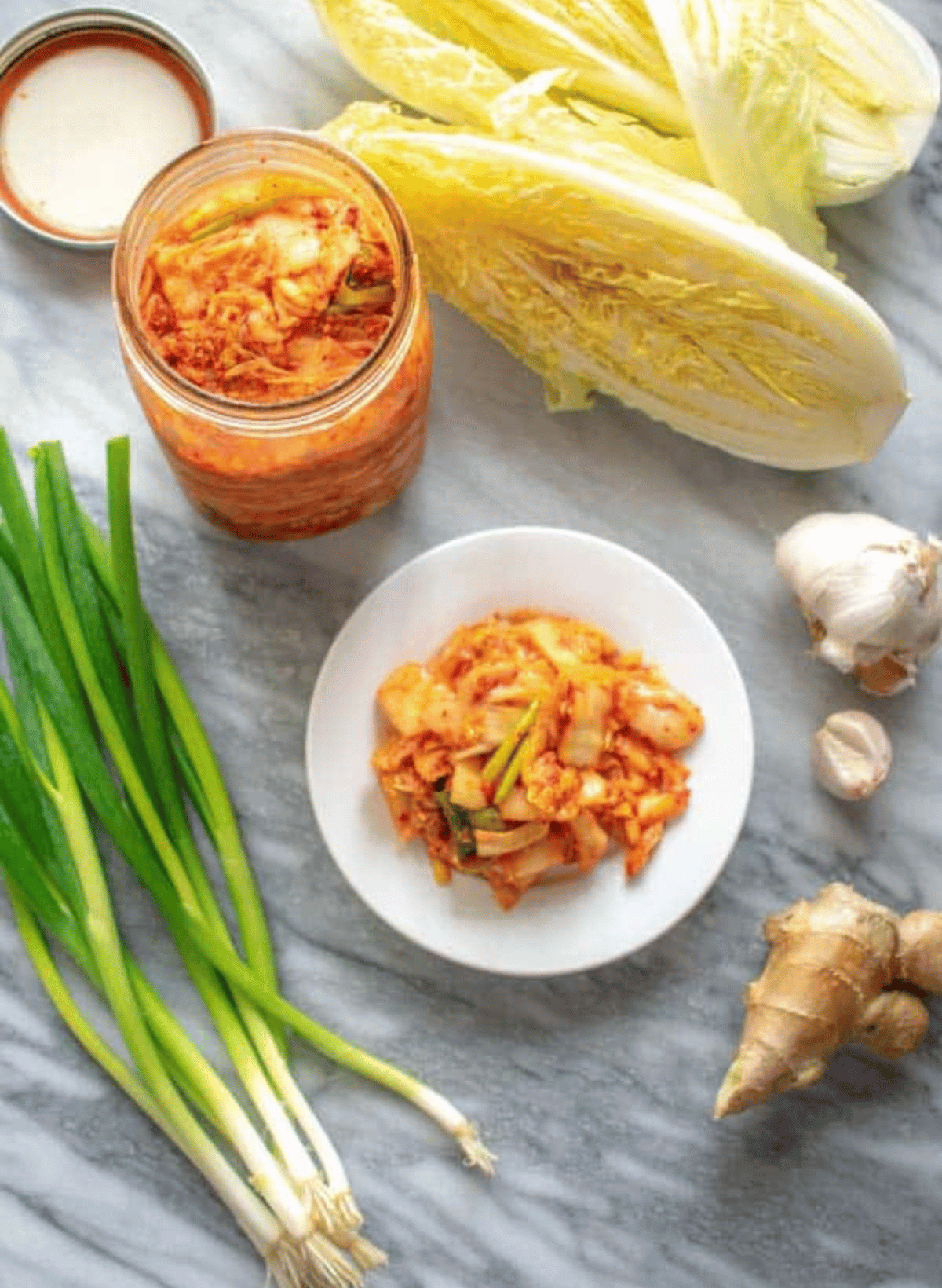 Red napa cabbage kimchi on a white round plate surrounded by kimchi ingredients and a mason jar of kimchi