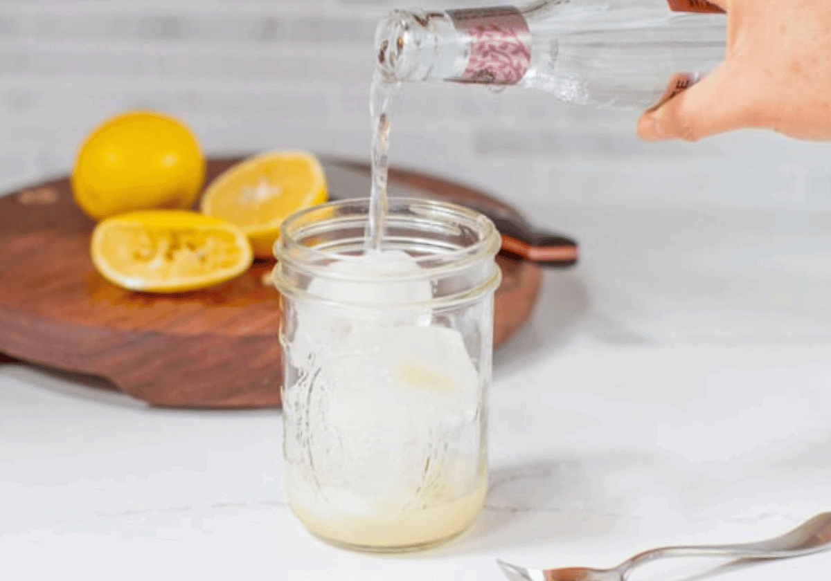 Glass bottle being poured into a mason jar with ice for Vietnamese lemon soda chanh with lemons and cutting board in the background