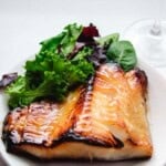 Nobu's Chilean Sea Bass on a white round plate with spring mix greens and a glass of red wine in the background