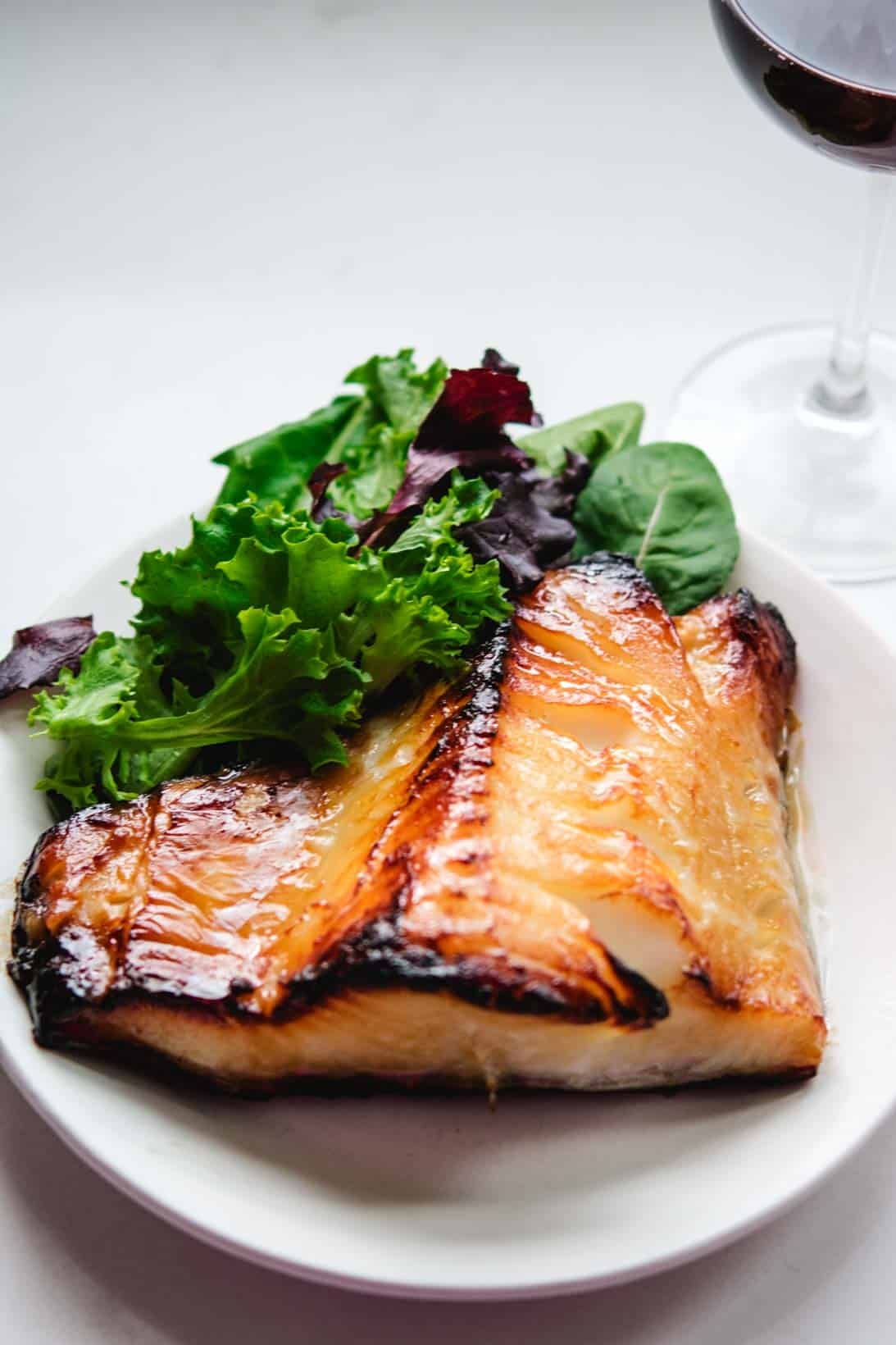 Nobu's Chilean Sea Bass on a white round plate with spring mix greens and a glass of red wine in the background