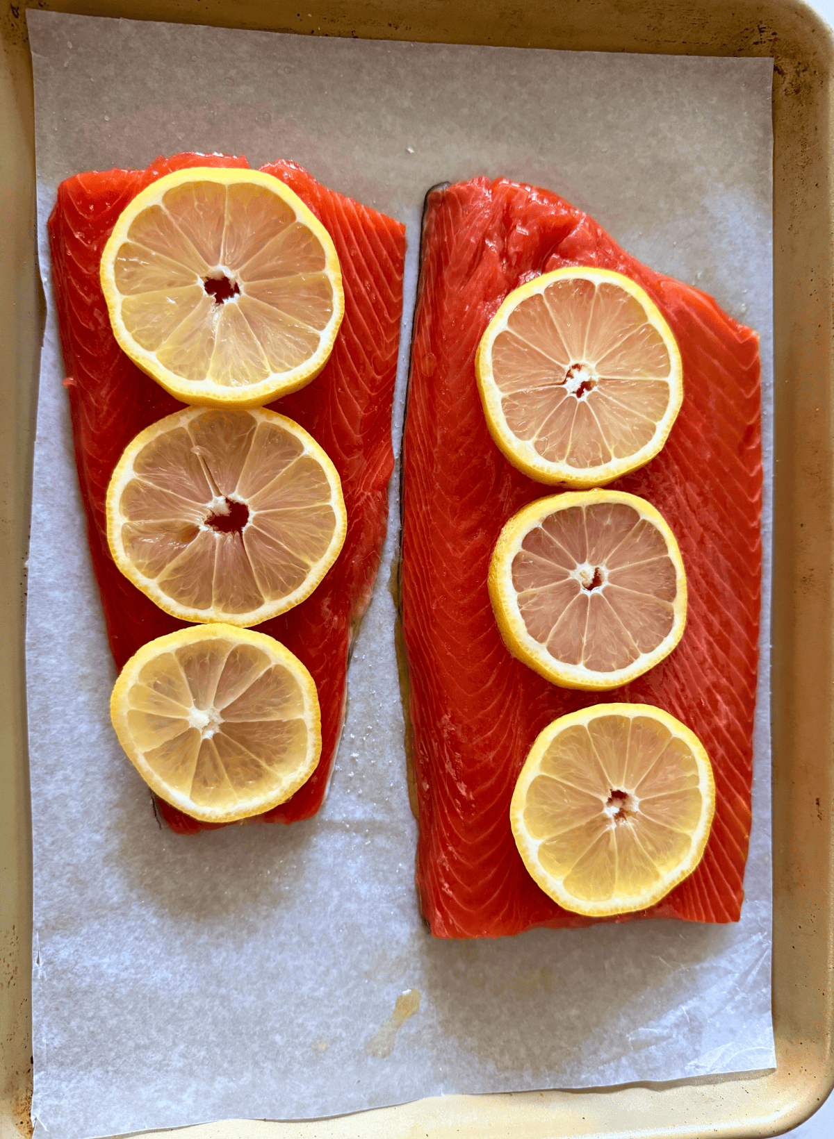 2 raw fillets of cooked slow roasted sockeye salmon with lemon slices on a parchment lined gold baking sheet