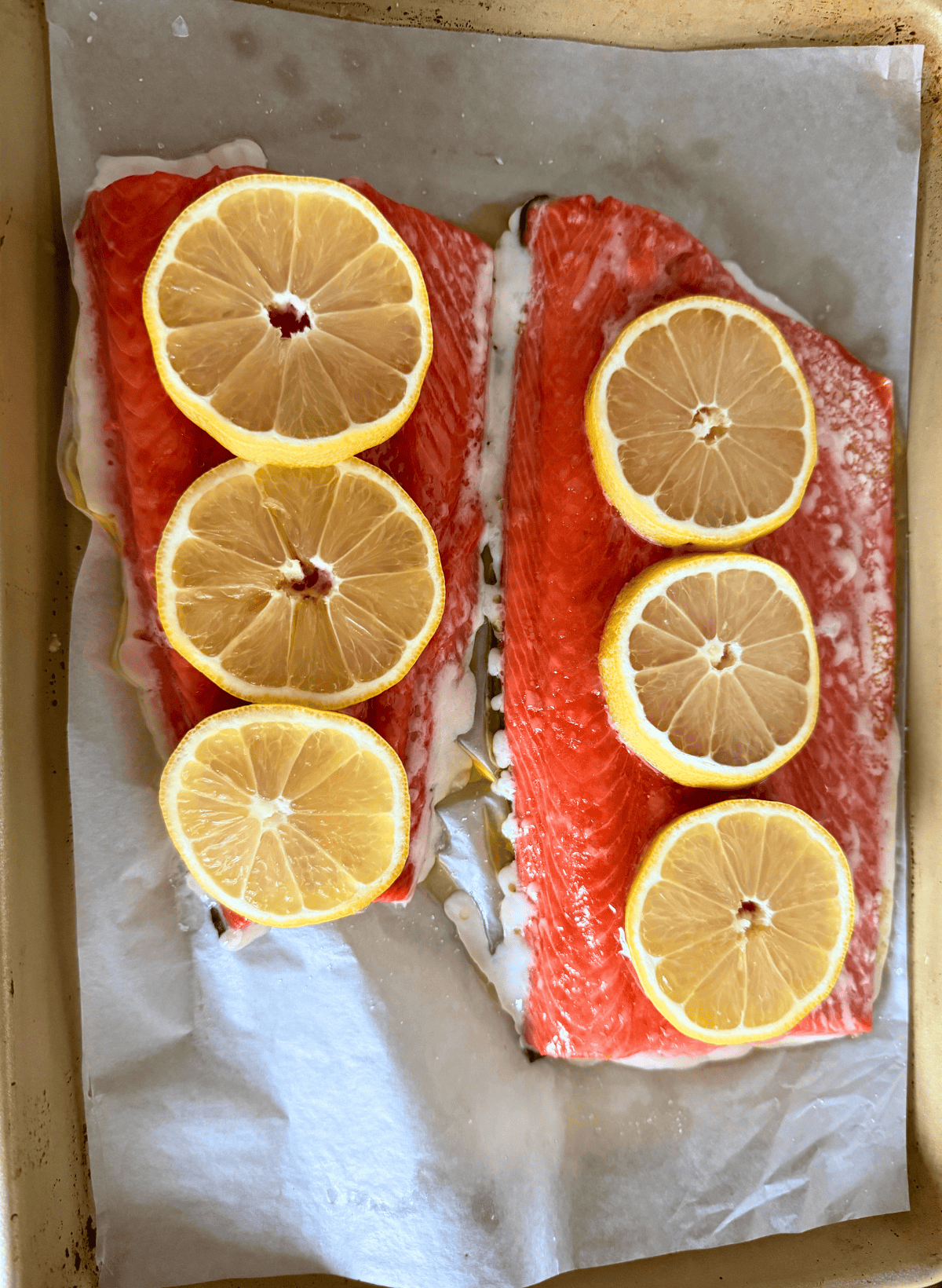 2 fillets of cooked slow roasted sockeye salmon with lemon slices on a parchment lined gold baking sheet