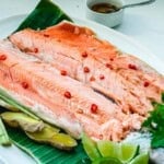 Cooked whole Coho salmon split open with bones removed on a banana leaf lined white platter and red chili garnish, lime wedges, ginger, and lemongrass