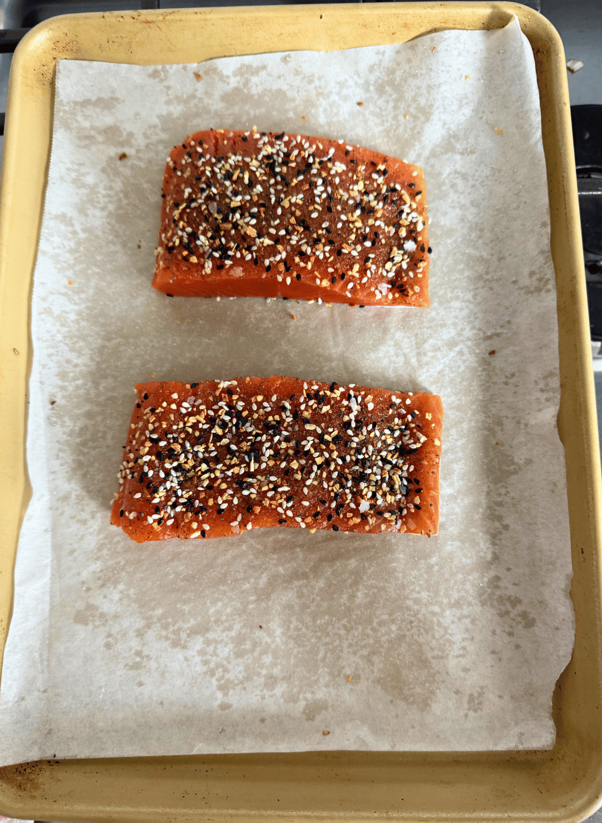 two fillets of raw coho salmon with everything bagel seasoning on a parchment lined gold baking sheet