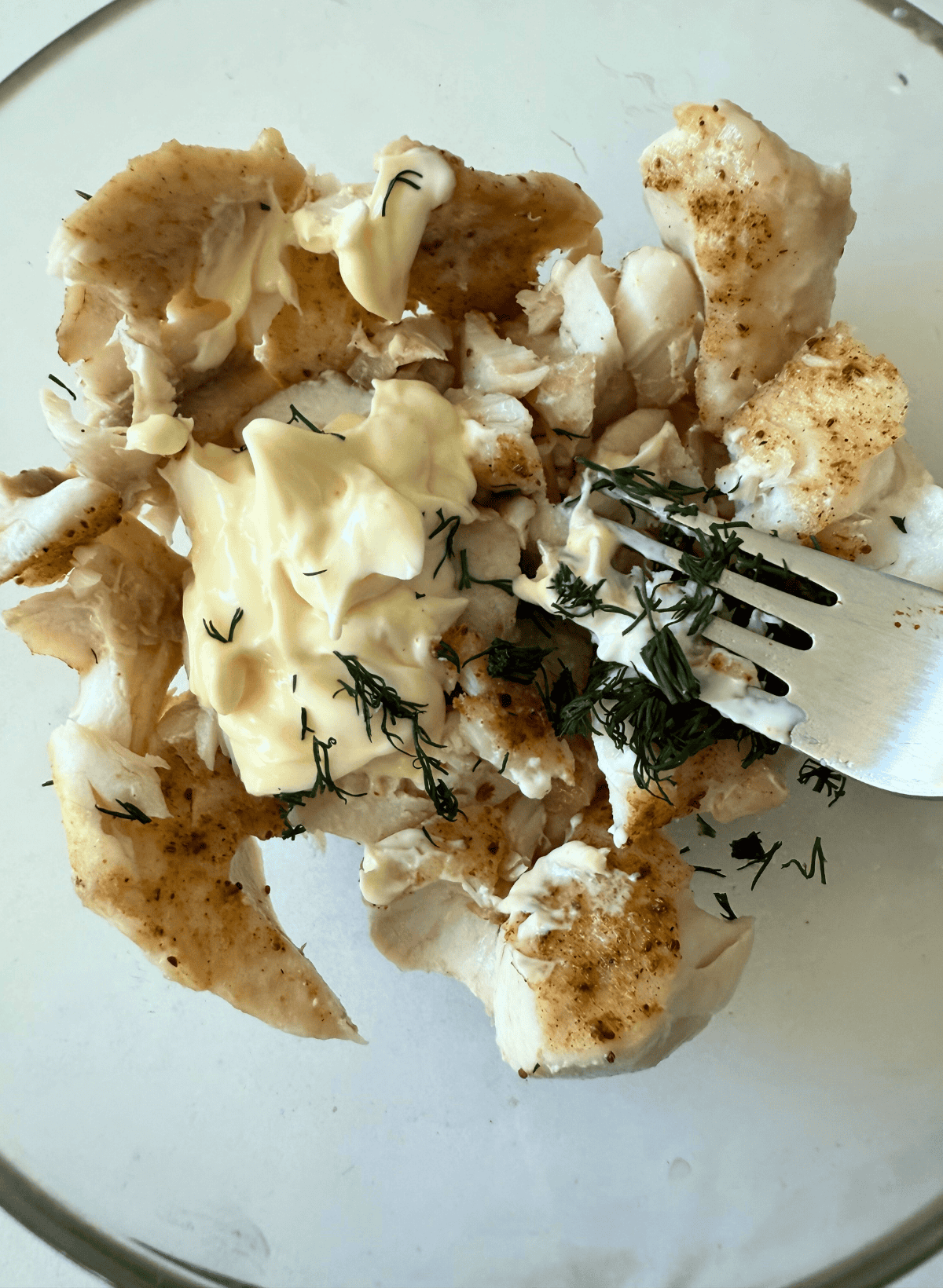 rockfish fillet being mixed with mayo and dill with a fork in a glass bowl